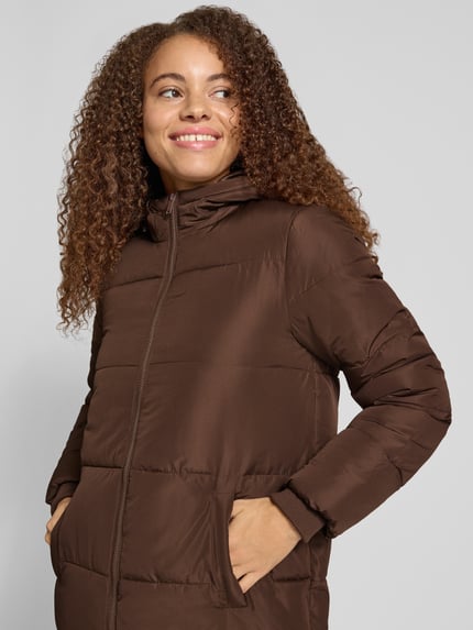 Frauenmodel trägt Puffer Coat mit Kapuze von PIECES, unifarben braun, Longline, Reißverschluss, Taschen, langarm, Ganzkörperansicht von vorne.