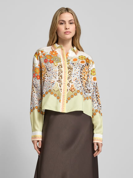 Modelka nosi Boxy Fit Bluse aus Satin im Allover-Look od Herzensangelegenheit, wzór kwiatowy, kołnierzyk koszulowy, jasne kolory, satynowa faktura, całkowity widok z przodu.