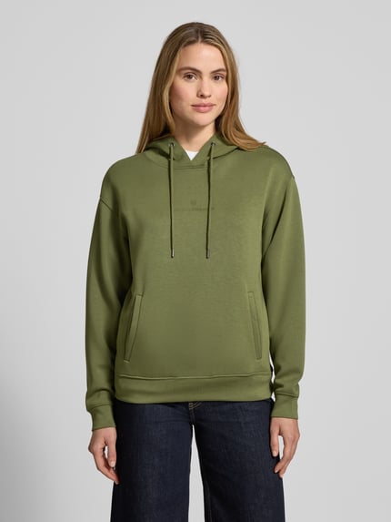 Frauenmodel trägt Hoodie aus Viskose-Mix mit Logo Modell 'IMA' von MSCH Copenhagen in Olivgrün, Kängurutaschen, Ganzkörperansicht von vorne.
