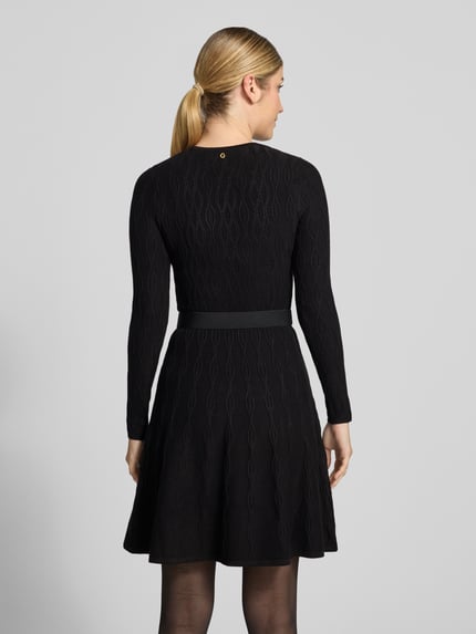 Männermodel trägt czarną sukienkę z dzianiny GUESS z mieszanki wiskozy z dodatkiem streczu, Linia a, Sukienka mini, Model 'YUKI', ścieg warkoczowy, okrągły dekolt, pasek, długość tyłu w rozmiarze S: 89 cm, szerokość w rozmiarze S: 32 cm, długość rękawów w rozmiarze S: 58 cm, widok całej sylwetki od tyłu.