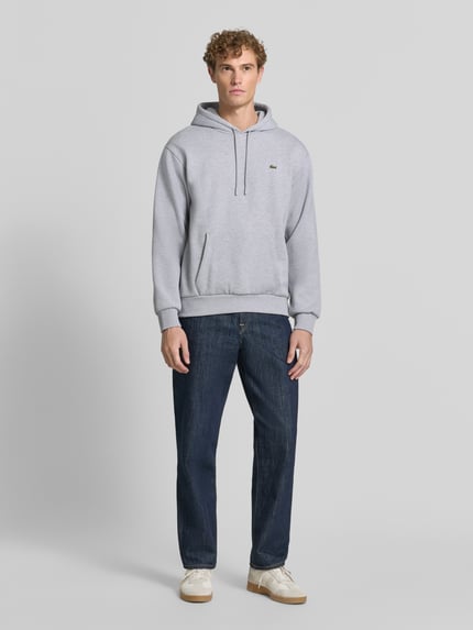 Männermodel trägt Relaxed Fit Hoodie aus Baumwoll-Mix von Lacoste in Hellgrau, kombiniert mit Jeans, Ganzkörperansicht von vorne. Herren Sweatshirt von Lacoste.