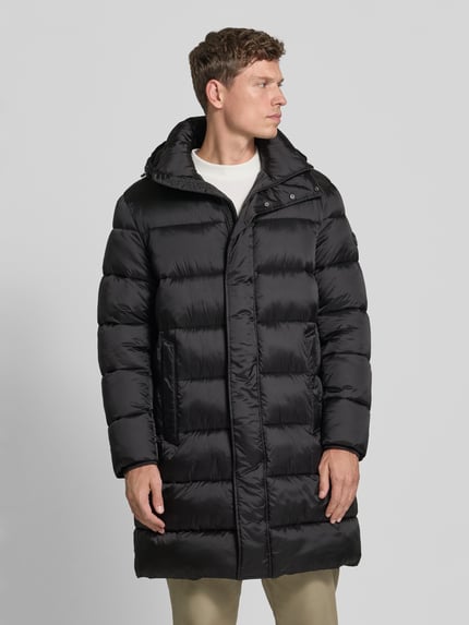 Männermodel trägt Pufferjacke mit Kapuze Modell 'JC-315Norman' von JOOP!, unifarben schwarz, Longline, mit Taschen und Logo-Patch, Ganzkörperansicht von vorne.