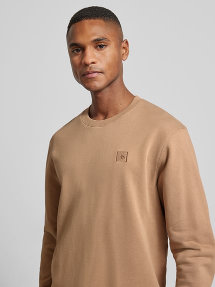Männermodel trägt Sweatshirt mit Label-Detail von Scotch & Soda in Beige, reine Baumwolle, lockerer Schnitt, Rundhalsausschnitt, Nahaufnahme Oberkörper.