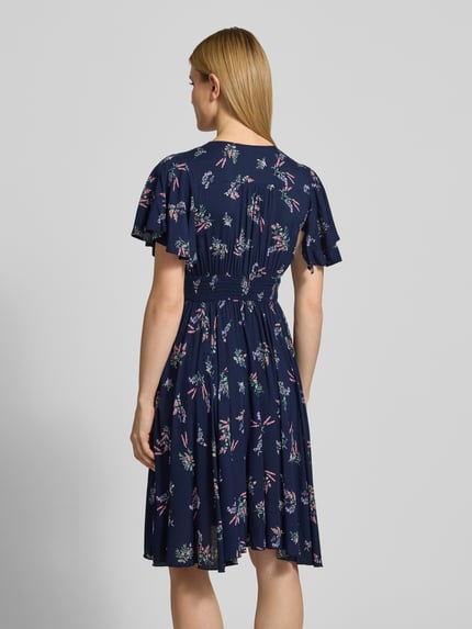 Frauenmodel trägt Knielanges Kleid aus Viskose mit V-Ausschnitt von Apricot in Dunkelblau mit Allover-Look, A-Linie, Knopfleiste, Elastisches Taillenband, Knielänge, Rückenlänge 104 cm, Brustweite 44 cm, Ärmellänge 21 cm, Ganzkörperansicht Rückansicht.