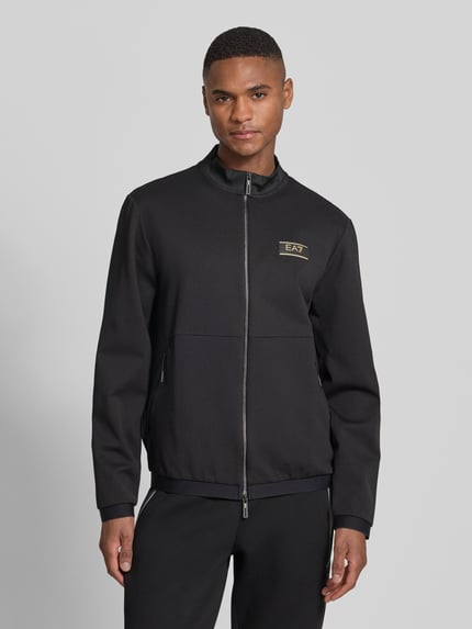 Männermodel trägt Sweatjacke mit Zweiwege-Reißverschluss von EA7 Emporio Armani, Schwarz, Label-Patch, Material-Mix, Ganzkörperansicht von vorne.