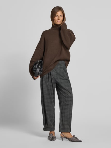 Frauenmodel trägt Wollpullover mit Rollkragen von Style Icon Collection in Braun, lockerer Schnitt aus reiner Wolle, gerippte Abschlüsse, dazu karierte Hose und graue Schuhe, Ganzkörperansicht von vorne.