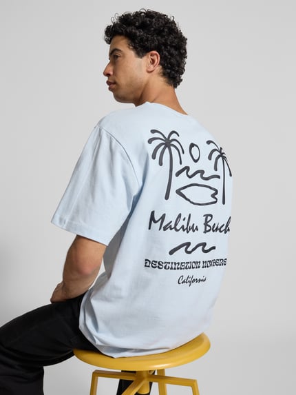 Męski model nosi Relaxed Fit T-Shirt aus reiner Baumwolle Modell 'MILO' od Only & Sons, kolor jasnoniebieski, print z motywem i napisem, widok całej sylwetki z tyłu.