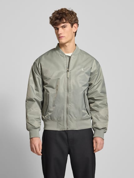 Męski model nosi Bomberjacke mit Label-Stitching od Calvin Klein Jeans, szaro-zielona, regular fit, długi rękaw, z przodu kieszenie, cała sylwetka z przodu.