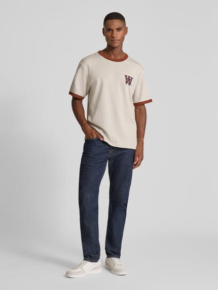 Männermodel trägt T-Shirt mit geripptem Rundhalsausschnitt Modell 'Parker' von Wood Wood in Beige mit Label-Print, Reine Baumwolle, Lockerer Schnitt, Rückenlänge 68 cm, Ganzkörperansicht von vorne.