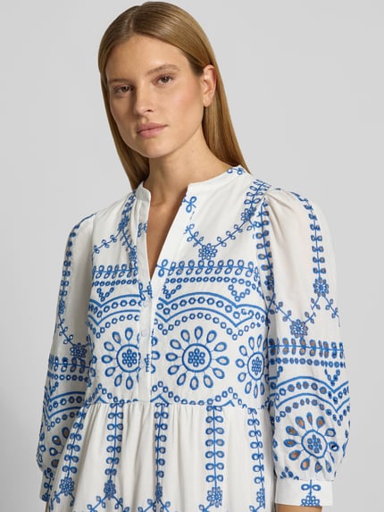 Vrouwenmodel draagt 'Tunikakleid mit Volantsaum und Lochstickerei' van Apricot, puur katoen, los model, serafinokraag, ajourwerk, korte knoopsluiting, volantzoom, midilengte, kleur wit met blauwe borduursels, detailopname van bovenlichaam.