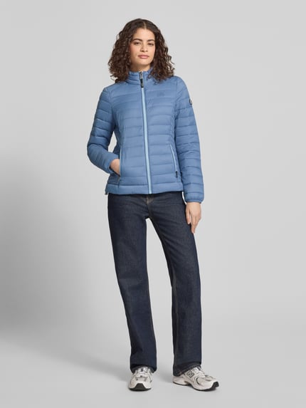Modelka kobieca ma na sobie jasnoniebieską Steppjacke mit Logo-Stitching Modell 'FUJI' od Superdry, prosty krój, stójka, zamek błyskawiczny, boczne kieszenie, długość tyłu w rozmiarze S: 62 cm, szerokość ramion w rozmiarze S: 40 cm, szerokość klatki piersiowej w rozmiarze S: 50 cm, długość rękawa w rozmiarze S: 69 cm, kolor jednolity, tekstura pikowana, pełny widok z przodu