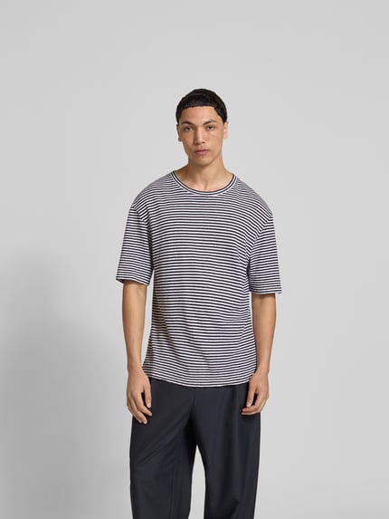 Męski model nosi Regular Fit T-Shirt aus Leinen-Mix von Officine Générale, pasiasty wzór, biało-granatowy, okrągły dekolt, półrękaw, len z bawełną, długość tyłu w rozmiarze M: 75 cm, krój regular fit, Ganzkörperansicht von vorne.