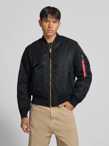 Männermodel trägt Bomberjacke mit Pattentaschen und Label-Applikation von Alpha Industries in Schwarz, Reißverschluss, Nahaufnahme Oberkörper. Regular Fit, Stehkragen, rote Label-Applikation am Ärmel, glatte Oberfläche, sichtbare Strukturdetails, Kontext: Rückenlänge 69 cm, Schulterbreite 44 cm, Brustweite 54 cm, Ärmellänge 64 cm.