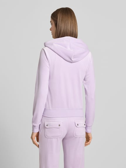 Frauenmodel trägt Slim Fit Sweatjacke aus Samt Modell 'Robertson' von Juicy Couture, elastyczna mieszanka materiałów, kaptur z tunel, zamek błyskawiczny, kieszeń kangurka, elastyczne wykończenia, kolor jasnofioletowy, akcenty z aksamitu, tekstura miękka, Ganzkörperansicht Rückansicht.