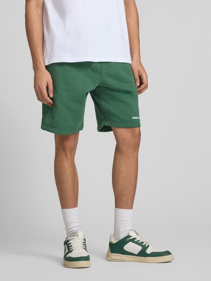 Männermodel trägt Shorts mit elastischem Bund und Logo-Stitching von Pegador aus Baumwoll-Mix in Grün, Loose-Fit, Nahaufnahme Unterkörper. Bundweite 38 cm, Innenbeinlänge 23 cm, französische Eingrifftaschen.