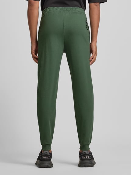 Männermodel trägt grüne Sweatpants mit Logo und elastischem Bund von EA7 Armani aus Baumwoll-Elasthan-Mix, Regular Fit, Ganzkörperansicht Rückansicht.