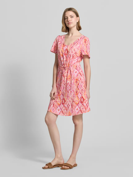 Frauenmodel trägt tailliertes Freizeitkleid aus reiner Viskose Modell 'EASY JOY' von Vero Moda mit Allover-Muster in Rosa und Orange, Mini-Länge, V-Ausschnitt, Kurzarm, glatte Textur, Ganzkörperansicht von vorne.