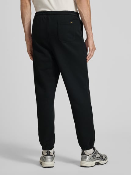 Männermodel trägt czarne spodnie dresowe Sweatpants mit Label-Print von Alpha Industries, mieszanka bawełny, luźny krój, elastyczny pas z tunelem, wpuszczane kieszenie boczne, nadruk z logo, Długość wewnętrzna nogawki w rozmiarze M: 44 cm, Ganzkörperansicht Rückansicht