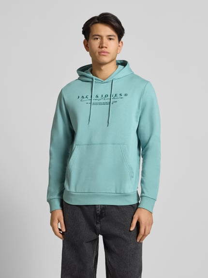 Männermodel trägt Hoodie mit Label-Print von Jack & Jones in Mint, Baumwoll-Mix, Känguru-Tasche, lockerer Schnitt, Ganzkörperansicht von vorne.