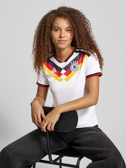 Vrouwenmodel draagt Trikot mit V-Ausschnitt Modell 'DFB' van adidas Sportswear, slim fit, label-stitching, wit met gekleurde details, nahaufnahme bovenlichaam.