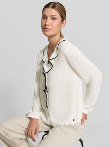 Frauenmodel trägt Regular Fit Bluse mit Volants von Marc Cain in Offwhite mit schwarzem Saum, V-Ausschnitt, Label-Detail, Nahaufnahme Oberkörper.