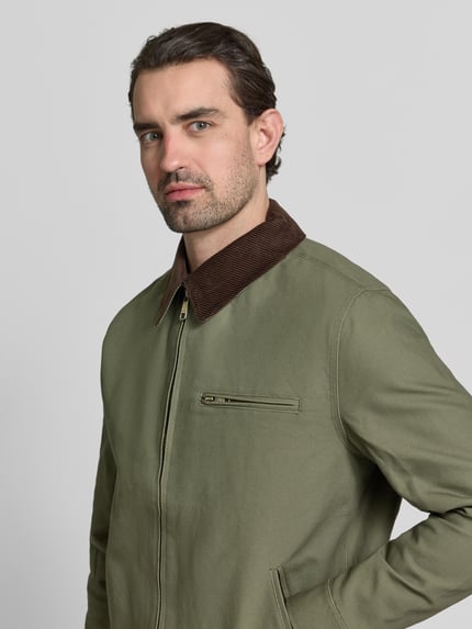 Męski model nosi Regular Fit Jacke mit Umlegekragen od MCNEAL, oliwkowa bawełna, kieszeń na piersi, zamek, zbliżenie górnej części ciała. Długość tyłu w rozmiarze M: 66 cm, szerokość ramion w rozmiarze M: 48 cm, szerokość klatki piersiowej w rozmiarze M: 60 cm, długość rękawa w rozmiarze M: 68 cm.