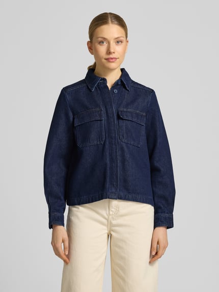 Frauenmodel trägt Jeansbluse mit Brusttaschen und Kentkragen von Stefanel, blaue Baumwoll-Mix-Struktur, Regular Fit, verdeckte Knopfleiste, Sportmanschetten, Ganzkörperansicht von vorne.