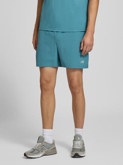 Männermodel trägt Shorts mit elastischem Bund und Logo von New Balance in Blau, Regular Fit, Nahaufnahme Unterkörper, Kontext: elastischer Bund, Tunnelzug, Logo, Bundweite M 39 cm, Innenbeinlänge 12 cm.