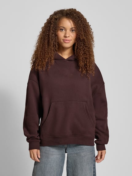 Frauenmodel trägt Hoodie mit rückseitigem Print Modell 'Good Karma Wave' von OH APRIL in Dunkelbraun aus Baumwoll-Mix, Oversized, Känguru-Tasche, Ganzkörperansicht von vorne.