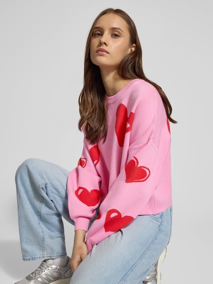 Frauenmodel trägt Oversized Strickpullover aus Viskose-Mix Modell 'SHYLA' von Noisy May, pink mit roten Herzen, Statement-Schriftzug, Ballonärmeln, O-Neck, Langarm, elastische Ärmelabschlüsse, Rückenlänge 44 cm, Ärmellänge 46 cm, Ganzkörperansicht von vorne.