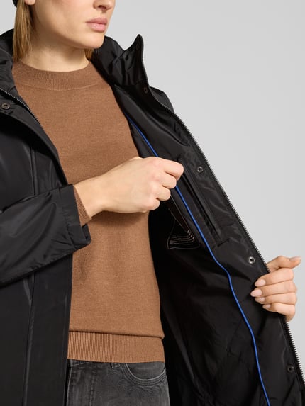 Kobiece modelka nosi Regular Fit Płaszcz z aplikacją z logo od NAUTGÅRD, stójka z detalem z logo, kaptur z tunelem, wpuszczane kieszenie, długość tyłu w rozmiarze S: 88 cm, szerokość ramion w rozmiarze S: 37 cm, szerokość klatki piersiowej w rozmiarze S: 48 cm, długość rękawów w rozmiarze S: 63 cm, kolor czarny, gładka tekstura, zbliżenie na górną część ciała