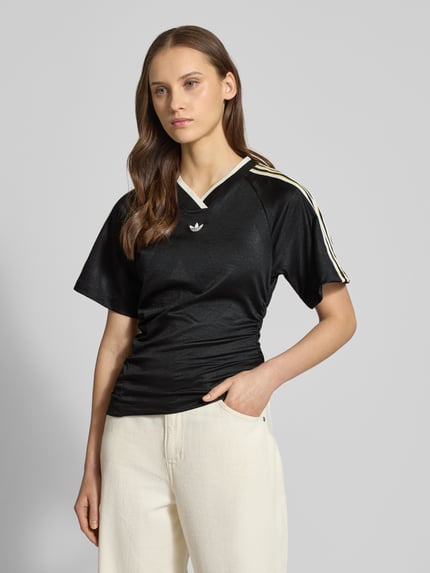 Frauenmodel trägt T-Shirt mit V-Ausschnitt von adidas Originals in Schwarz mit Label-Details, asymmetrisch, Kurzarm, Ganzkörperansicht von vorne.