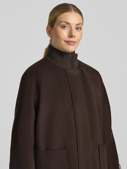 Frauenmodel trägt Mantel aus Woll-Mix mit gestricktem Stehkragen von COMMA in dunklem Braun, sichtbare Struktur, Nahaufnahme Oberkörper. Straight Fit, Viskose-Anteil, Reiß- und Druckknopfverschluss, seitliche Taschen, gestrickte Ärmelabschlüsse.