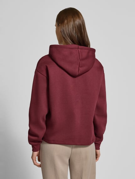 Modelka kobieca ma na sobie Regular Fit Hoodie aus Baumwoll-Mix Modell 'CHILLI' od Pieces, kolor bordowy, gładki, tkanina sweat, krój regular fit, kaptur z troczkiem, długie rękawy z prążkowanym mankietem, widok całej sylwetki od tyłu.