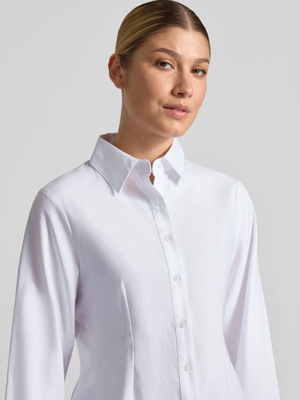 Frauenmodel trägt Oversized Fit Hemdbluse aus Baumwoll-Mix mit Ziernähten von Neo Noir, weiße Farbe, glatte Textur, Nahaufnahme Oberkörper. Kontext: Damen Hemdbluse, durchgängige Knopfleiste, Hemdblusenkragen, langärmelig, 1-Knopf-Manschetten, Rückenlänge 55 cm, Ärmellänge 61 cm.