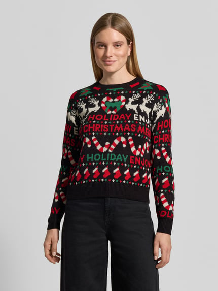 Frauenmodel trägt Pullover mit Weihnachtsmotiv-Frontprint von VERO MODA, mehrfarbig, grafisches Allover-Muster, Strickoptik, Ganzkörperansicht von vorne.