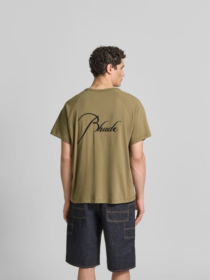 Męski model nosi Regular Fit T-Shirt aus reiner Baumwolle od RHUDE, kolor khaki, wyhaftowane logo, raglanowe rękawy, okrągły dekolt, długość tyłu w rozmiarze M: 69 cm, widok całej sylwetki z tyłu.