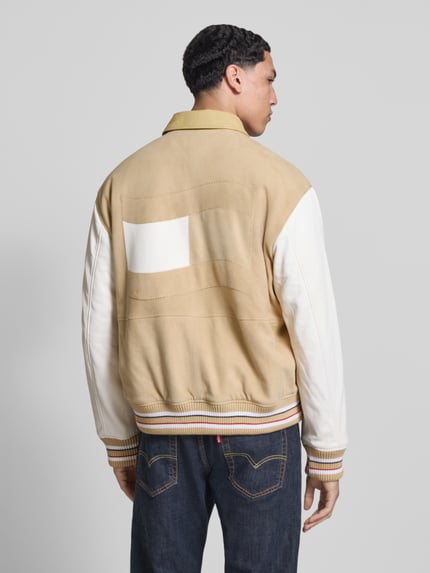 Männermodel trägt Relaxed Fit College-Jacke aus echtem Leder von Tommy Jeans in Beige und Weiß, Veloursleder-Optik, Ganzkörperansicht Rückansicht.
