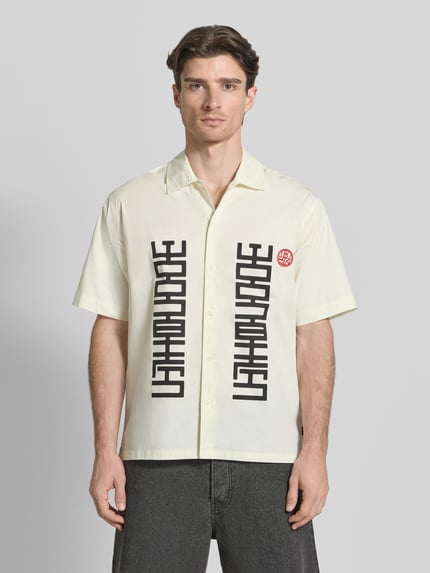 Männermodel trägt Regular Fit Freizeithemd mit Label-Print Modell 'Banks' von WOODBIRD, cremefarben mit schwarzen Details, ganzkörperansicht von vorne, context: heren vrijetijdsoverhemd, regular fit, label-print, kentkraag, ruglengte maat M 76 cm, schouderbreedte 52 cm, mouwlengte 28 cm