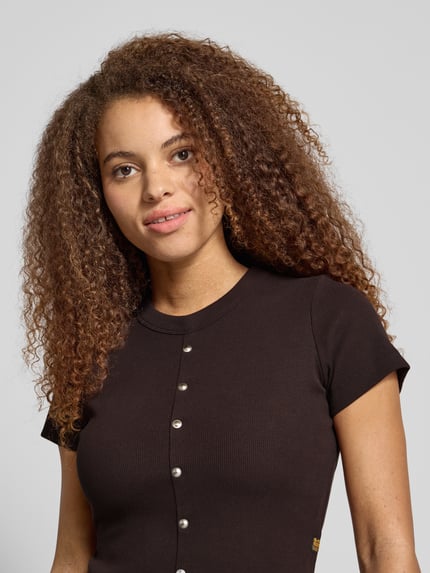 Młodzieżowy modelka nosi czarne Cropped T-Shirt mit Knopfleiste Modell 'Heritage' od G-Star Raw, bawełna z elastanem, krótki rękaw, faktura prążkowana, okrągły dekolt, metalowe guziki, widoczne logo, zbliżenie na górną część ciała.