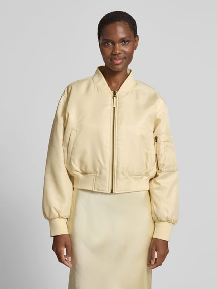 Frauenmodel trägt Bomberjacke mit Stehkragen von Anine Bing in hellem Beige, glänzende Textur, Regular-Fit, Reißverschluss, Eingrifftaschen, elastische Abschlüsse, Rückenlänge bei Größe S 50 cm, Brustweite 60 cm, Ärmellänge 58 cm, Ganzkörperansicht von vorne.