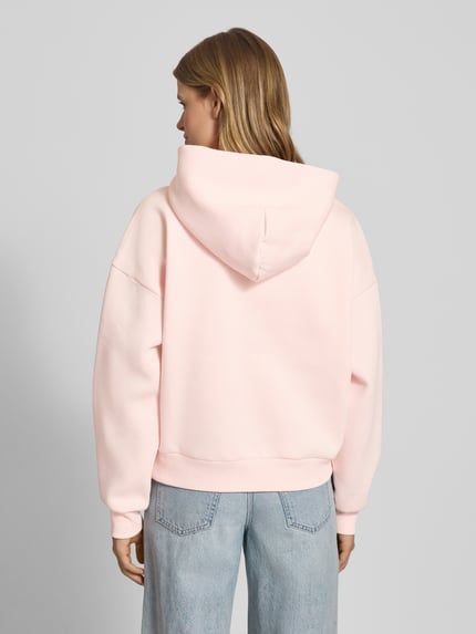 Frauenmodel trägt Oversized Hoodie mit Kapuze von Review in Hellrosa, Baumwoll-Mix, gerippte Abschlüsse, Känguru-Tasche, Ganzkörperansicht Rückansicht.