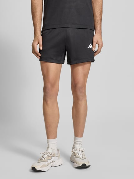 Mężczyzna nosi Straight Fit Shorts mit Logo-Print in Mesh-Optik von adidas Training, czarne, siateczkowa faktura, elastyczny pas, szerokość w rozmiarze M: 39 cm, długość wewnętrzna nogawki: 12 cm, materiał matowy, Ganzkörperansicht z przodu.