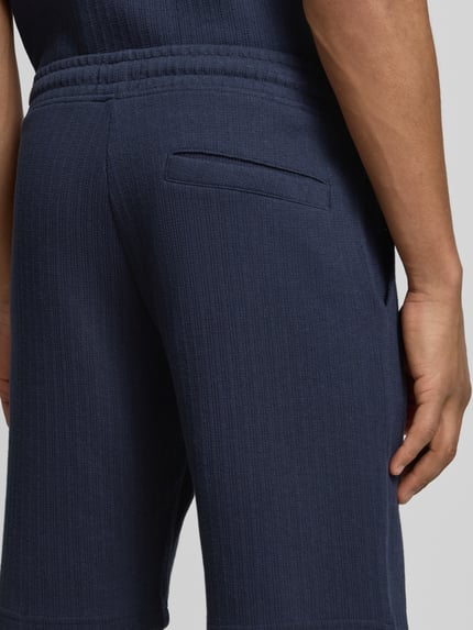 Mężczyzna nosi Regular Fit Shorts mit Tunnelzug von MCNEAL z mieszanki bawełny z dodatkiem streczu, wpuszczane kieszenie, kieszeń z tyłu, kolor granatowy, tekstura prążkowana, długość wewnętrzna nogawki 23 cm, szerokość w rozmiarze M 43 cm, detal tylnej części krótkich spodni, zbliżenie tylnej części.