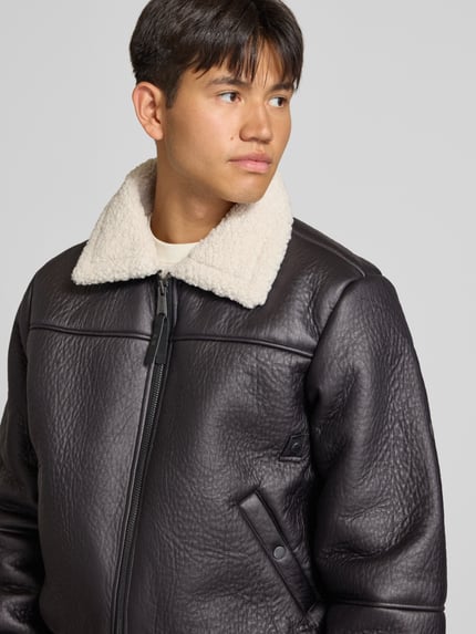 Männermodel trägt Bomberjacke mit Kentkragen und gerippten Abschlüssen von Alpha Industries in Schwarz, Kunstleder-Optik, Nahaufnahme Oberkörper. Regular Fit, Teddyfell am Kragen, Reißverschluss, Eingrifftaschen mit Druckknopf, Rückenlänge 66 cm, Schulterbreite 48 cm, Brustweite 55 cm, Ärmellänge 70 cm.