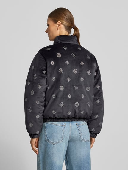 Młody modelka nosi czarną kurtkę GUESS Activewear Jacke im Allover-Look Modell 'AMALIA' z ozdobnymi kamieniami, krój regular fit, materiał mieszany z dodatkiem stretchu, stójka, zamek, tekstura gładka z nadrukami, widoczne szwy, jeansy, zdjęcie całej sylwetki od tyłu.