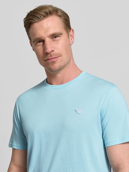 Männermodel trägt T-Shirt mit Logo-Patch und Rundhalsausschnitt von Emporio Armani in Hellblau, Reine Baumwolle, Modern Fit, Nahaufnahme Oberkörper. Rückenlänge bei Größe M: 69 cm.
