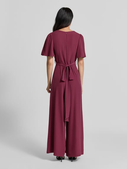 Frauenmodel trägt Jumpsuit mit Stoffgürtel von Rinascimento in Bordeaux, glatter Stoff, taillierte Passform, Ganzkörperansicht Rückansicht. Damen Jumpsuit von Rinascimento, Material-Mix mit Stretch-Anteil, V-Ausschnitt, Kurzarm, Raffungen, Stoffgürtel, Maße bei Größe S.