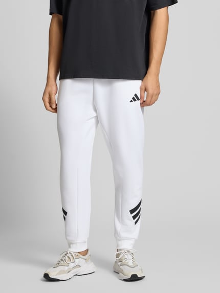 Männermodel trägt Sweatpants mit Logo und elastischem Bund von adidas Sportswear, weiß, schwarzes Logo, Material-Mix, Regular-Fit, Ganzkörperansicht von vorne.