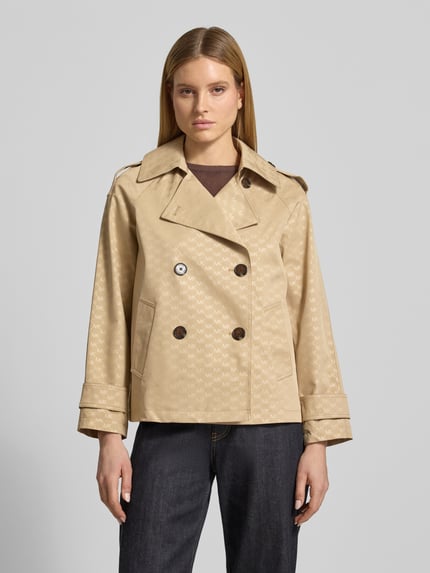 Vrouwenmodel draagt trenchcoat met logomotief van MICHAEL Michael Kors in regular fit, met dubbele rij knopen, reverskraag, mouwsluitingen; ruglengte maat S 65 cm, schouderbreedte 48 cm, borstwijdte 53 cm, mouwlengte 57 cm. Ganzekörperaanzicht van voren.
